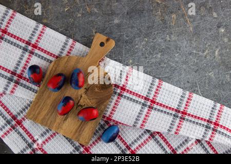 Mit einem Pinsel bemalte Eier in roten und blauen Farben liegen auf einem kleinen Holzbrett, auf einem Waffeltuch auf einem alten dunklen Marmor. Draufsicht, viel Platz Stockfoto