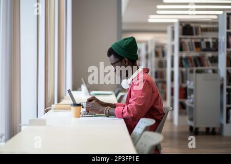 Fokussierter afroamerikanischer Student, der am Schreibtisch mit einem Laptop sitzt und eine Online-Bibliotheksdatenbank verwendet Stockfoto