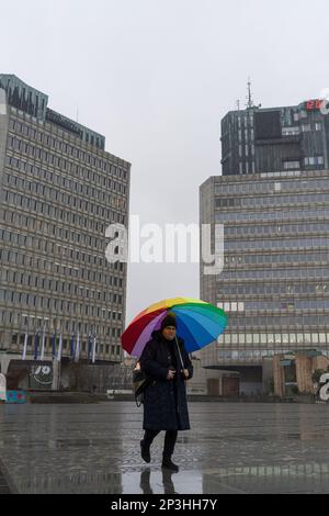 12 26 2022, Ljubljana, Slowenien: Eine Frau mit einem bunten Regenschirm, die den Platz der Republik/Trg republike hinuntergeht, eingerahmt von Ljubljanska Banka und TR3 bis Stockfoto