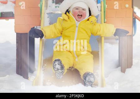 Der kleine Junge fährt auf einer Rutsche und spielt auf einem Winterspielplatz. Ein Kind in einem gelben Overall auf einer Kinderrutsche im Schnee. Ein Kind im Alter von einem Jahr Stockfoto