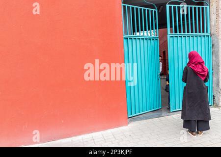 Eine Dame in Schwarz mit einem burgunderroten Schal, die vor einer blau-grünen eisernen Tür wartet, an einer roten Wand. Brüssel. Stockfoto