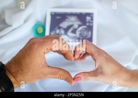Schwangere Frau und Ehemann halten Hand zusammen, halten Ultraschallbild und machen Hand-Herz-Geste. Erwartungen an die Schwangerschaft von Kindern und Frauen Stockfoto