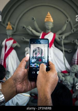 Ein Mann, der auf der Kolkata Book Fair ein Foto von Göttin Durga in Mobile macht. Selektiver Fokus auf Mobilgeräte mit anderen verschwommenen Bildern. Stockfoto