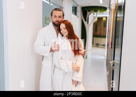 Porträt eines attraktiven jungen Paares, das verliebt ist, einen weißen Bademantel trägt, sich umarmt, mit einem Lächeln in die Kamera schaut und sich im Spa-Salon entspannen kann. Stockfoto