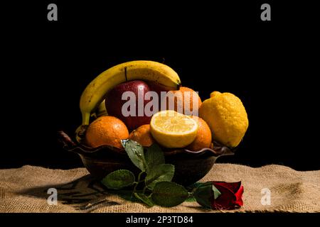 Stillleben von Früchten Bananen Orangen Zitronen Äpfel auf einem Tontopf auf dunklem Hintergrund Stockfoto