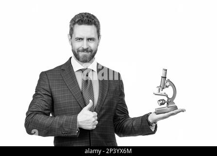 Ein gutaussehender Geschäftsmann im Geschäftsanzug, der ein modernes Mikroskop auf weißem Boden präsentiert, zeigt Daumen hoch, Wissenschaft und Wirtschaft. Stockfoto