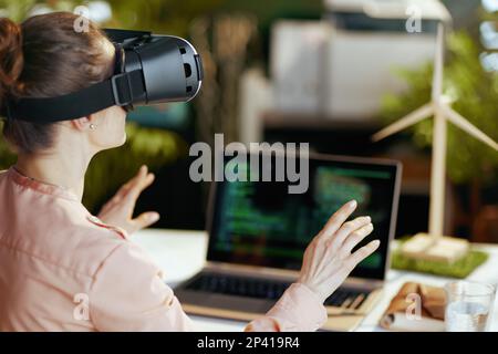 Moderne Frau mit Notebook und Virtual-Reality-Headset im modernen Büro von hinten gesehen. Stockfoto