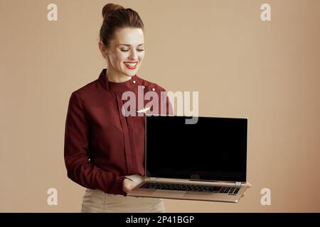 Lächelnde, moderne weibliche Stewardess auf beigem Hintergrund mit leerem Laptop-Bildschirm. Stockfoto