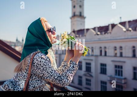 Stilvolle junge Frau mit grünem Retro-Schal und Sonnenbrille, die nach Frühlingsblumen riecht. Klassische Outdoor-Mode im Vintage-Stil Stockfoto