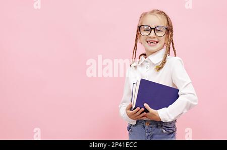 Ein bezauberndes Schulmädchen mit lustigen Zöpfen auf dem Kopf, das Lehrbücher in den Händen hält, in der Kamera lächelt und die Brille mit dem Finger auf pinkfarbenen Hintergrund fixiert, bereitet sich darauf vor Stockfoto