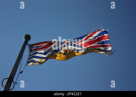 Ein Blick aus nächster Nähe auf eine Provinzflagge von British Columbia (BC), die an einem windigen Tag von einem Fahnenmast aus fliegt. Die Flagge ist hintergrundbeleuchtet und am Himmel ausgefranst Stockfoto