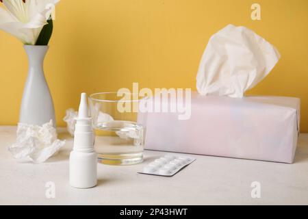 Nasentropfen mit Pillen, Taschentüchern, Glas Wasser und Blume auf dem Tisch nahe der gelben Wand. Saisonales Allergiekonzept Stockfoto