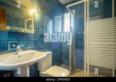 Badezimmer mit Spiegel an der Wand mit Glasregal, Porzellanwaschbecken, quadratischer weißer Duschkabine und superalten blauen Fliesen Stockfoto