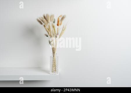 Kleine geschnitzte Glasvase mit Stacheln und getrockneten Pflanzenzweigen, einige auf einem weißen Holzregal und einer weißen Wand gefärbt Stockfoto