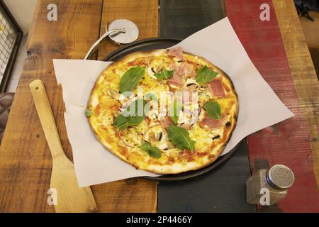 Tolle italienische Pizza mit dünner Kruste mit Pilzen, Prosciutto und viel Basilikum auf einem Holztisch Stockfoto