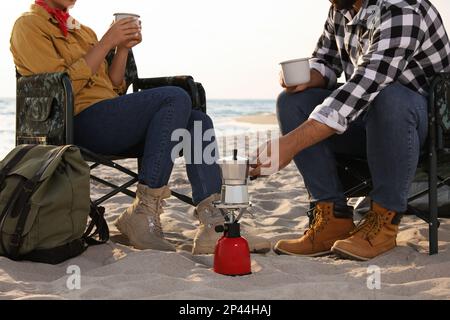 Ein Paar ruht sich am Strand in Campingstühlen aus und schließt. Ich mache Kaffee Stockfoto