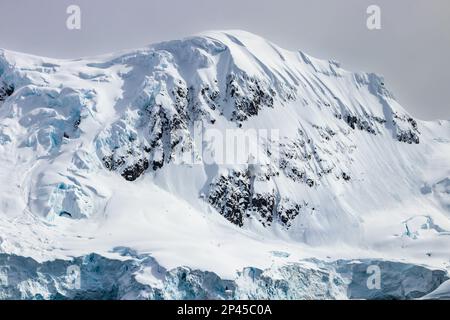 Schneebedeckter Berggipfel, antarktische Halbinsel. Felsen auf schneebedeckten Hängen. Blaues Eis sichtbar. Wolkiger Himmel im Hintergrund. Stockfoto