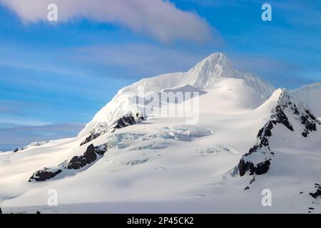 Schneebedeckte Bergspitze, am frühen Morgen, antarktische Halbinsel. Felsen auf schneebedeckten Hängen. Blaues Eis sichtbar. Blauer Himmel, Wolken im Hintergrund. Stockfoto