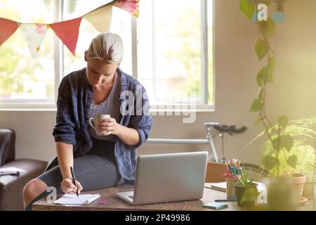 Lassen Sie den kreativen Prozess beginnen. Eine junge Frau, die Dinge auf einen Notizblock schreibt, während sie auf ihrem Schreibtisch sitzt und Kaffee trinkt. Stockfoto