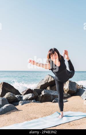 Glückliche Frau in Sportbekleidung balanciert auf dem Bein in Natarajasana. Sie sitzt auf einer Matte, während sie an sonnigen Tagen Yoga an der Sandküste in der Nähe des Meeres macht und in die Kamera schaut Stockfoto