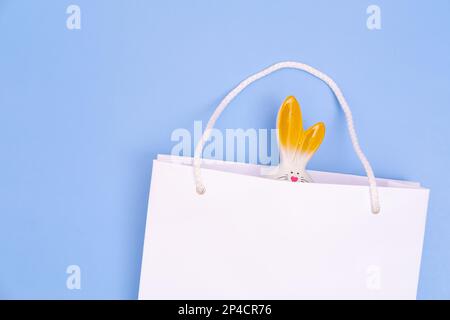 Ostern einkaufen. Weiße Einkaufstasche aus Papier mit weißem Häschen auf blauem Hintergrund. Konzept Ostergeschenke, Vorbereitung, Weihnachtsüberraschung. Stockfoto