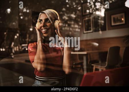 Lächelnde Frau, während sie mit Kopfhörern in einem Café Musik hört. Stockfoto
