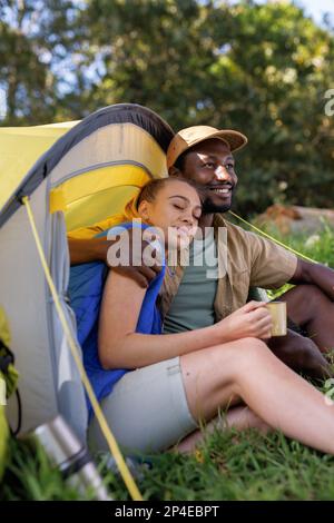 Vertikal eines glücklichen, vielfältigen Paares im Wald, sitzen am Zelt, trinken Kaffee, umarmen, Kopierraum Stockfoto