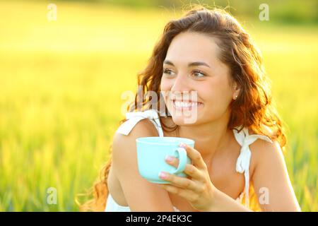 Glückliche Frau, die bei Sonnenuntergang eine Kaffeetasse auf einem wunderschönen Feld hält Stockfoto