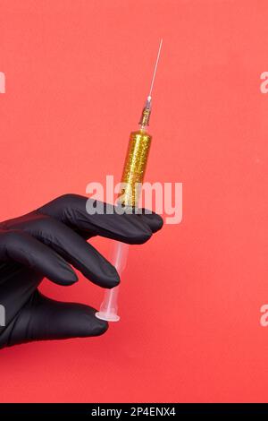 Handgehaltene Spritze gefüllt mit gelbem Glitzer. Vertikale Aufnahme auf rotem Hintergrund. Schwarzer Latexhandschuh. Stockfoto