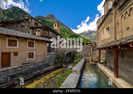 Der Alpenfluss fließt unter der mittelalterlichen Steinbrücke zwischen alten Häusern als Berge im Hintergrund im kleinen Dorf Chianale in Piedmont, Norditalien. Stockfoto