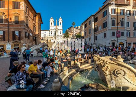 Menschen sitzen um die Fontana della Barcaccia (Brunnen des Langboots) auf der Piazza di Spagna als spanische Treppe im Hintergrund in Rom, Italien. Stockfoto