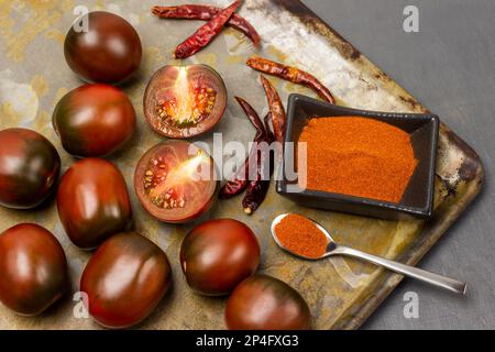 Gemahlener Pfeffer in einer Schüssel. Trockene Paprika-Hülsen. Frische Tomaten auf einem rostigen Tablett. Flach verlegt. Grauer Hintergrund. Stockfoto