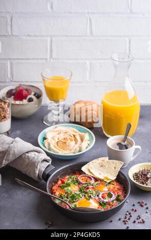 Großes, gesundes Frühstück mit nahöstlichem und europäischem Essen. Shakshuka Eier, Pita, Hummus, Hüttenkäse, Obst, Joghurt, Müsli, Croissant Stockfoto