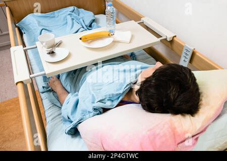 Kranke Frau und Tisch mit Geschirr und Banane auf dem elektrischen Krankenhausbett zu Hause Stockfoto