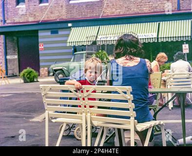 Ein historisches Bild aus dem Jahr 1981 von einem Kind, das vor der Kamera sitzt, und seiner Mutter, die an einem Tisch im Birkenhead Point Shopping Centre sitzt, das nur vier Jahre zuvor aus einer alten Reifenfabrik umgebaut wurde. Im Hintergrund befindet sich ein Gebäude mit gestreiften Jalousien aus Segeltuch und einem Toyota aus den frühen 1970er Jahren, der ute abholt Stockfoto