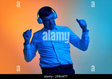 Porträt eines erwachsenen Mannes mittleren Alters, der Musik über Kopfhörer hört und über einen abgestuften blauen orangefarbenen Hintergrund in Neonlicht tanzt. Konzept von Stockfoto