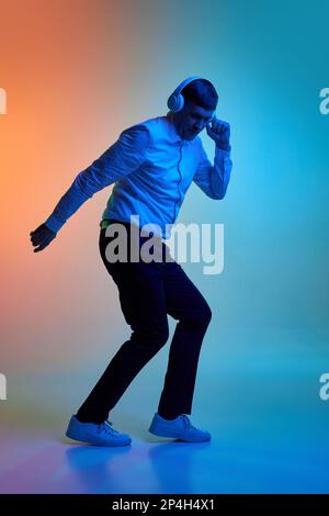 Porträt eines erwachsenen Mannes mittleren Alters, der Musik über Kopfhörer hört und über einen abgestuften blauen orangefarbenen Hintergrund in Neonlicht tanzt Stockfoto