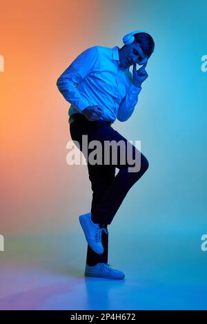 Porträt eines erwachsenen Mannes mittleren Alters, der Musik über Kopfhörer hört und über einen abgestuften blauen orangefarbenen Hintergrund in Neonlicht tanzt Stockfoto