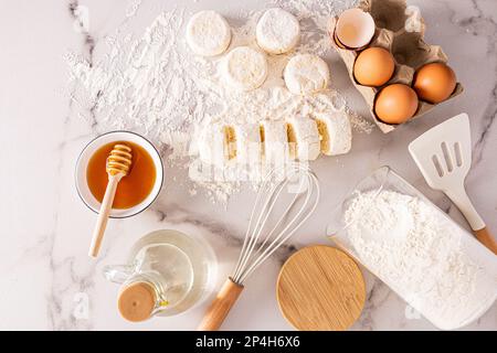 Blick von oben auf frisch zubereitete Käsekuchen und Zutaten zum Kochen. Eier, Honig, Butter in einer Flasche. Köstliches Frühstück Stockfoto