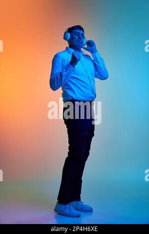 Porträt des positiven Mannes mittleren Alters in voller Länge, der Musik über Kopfhörer hört und über einen abgestuften blauorangefarbenen Hintergrund in Neonlicht tanzt Stockfoto