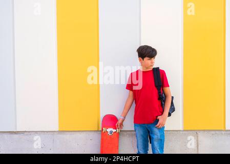 Ein Junge steht an der Wand und hält ein Skateboard, während er wegschaut Stockfoto