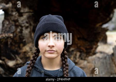 Teenager mit Zöpfen und Beanie steht vor dem gefallenen Baum Stockfoto