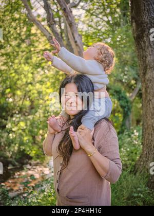 Die Tochter sitzt auf den Schultern der Mutter in der Nähe des Bachs in einer bewaldeten Umgebung Stockfoto