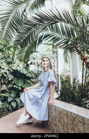Eine braune Frau in einem blauen Kleid sitzt unter einer Palme in einem Gewächshaus Stockfoto