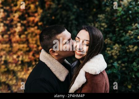 Ein junger Mann küsst eine lächelnde Frau auf der Wange gegen einen Park Stockfoto