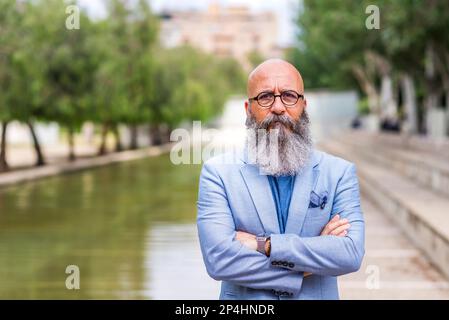 Glatze mittleren Alters, bärtig stehend, mit gekreuzten Armen posiert im Park Stockfoto