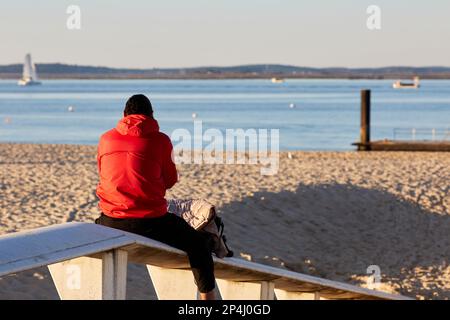 Ein junger Mann, der auf einer Bank sitzt, blickt aufs Meer Stockfoto