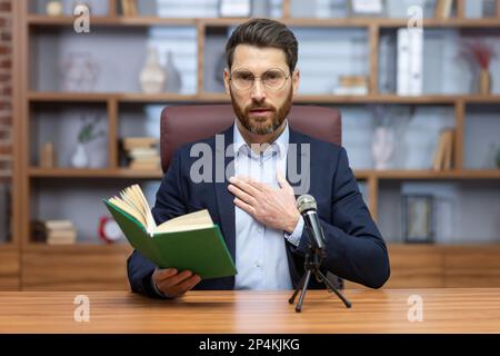Älterer Pastor liest online bibelbuch, Mann schaut in die Kamera, sitzt im Heimbüro und nimmt Hörbuch online auf. Stockfoto