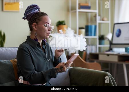 Junge Geschäftsfrau, die mit dem Handy scrollt und elektronische Zigaretten raucht, während sie in der Pause auf der Couch im Heimbüro sitzt Stockfoto