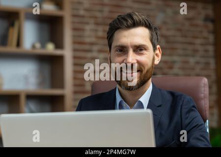 Nahaufnahme eines erfolgreichen, erwachsenen Geschäftsmanns im Heimbüro, bärtigen Mannes bei der Arbeit mit einem lächelnden Laptop und einem Blick auf die Kamera, der mit der Fernarbeit zufrieden ist. Stockfoto